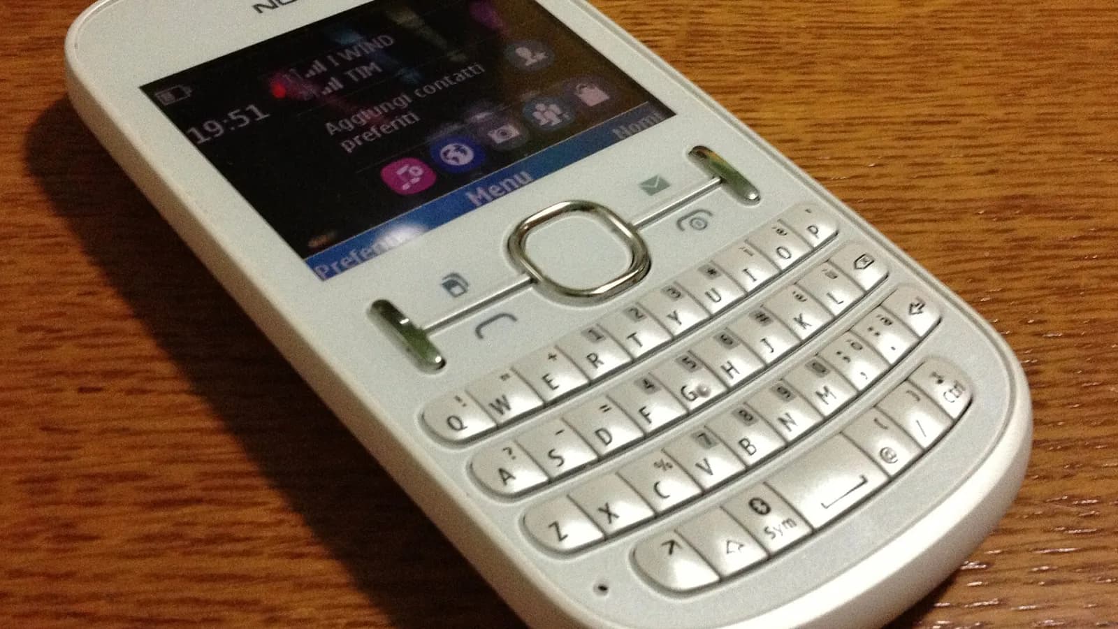 Telefon celular Nokia Asha 200 me ngjyrë të bardhë dhe tastierë QWERTY mbi një sipërfaqe druri.