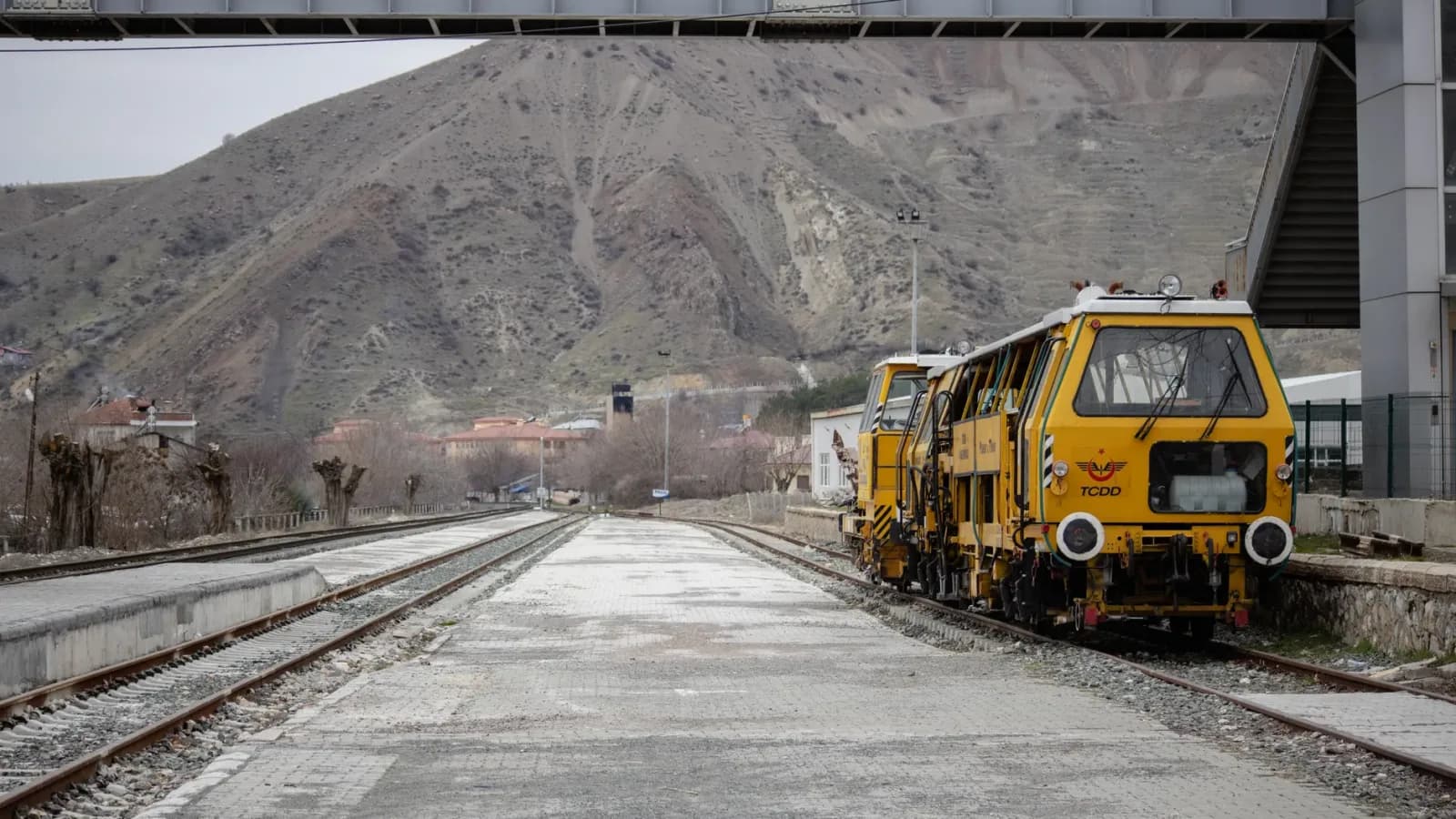 Makineri e verdhe per mirembajtjen e shinave hekurudhore e parkuar ne nje stacion treni me male ne sfond.