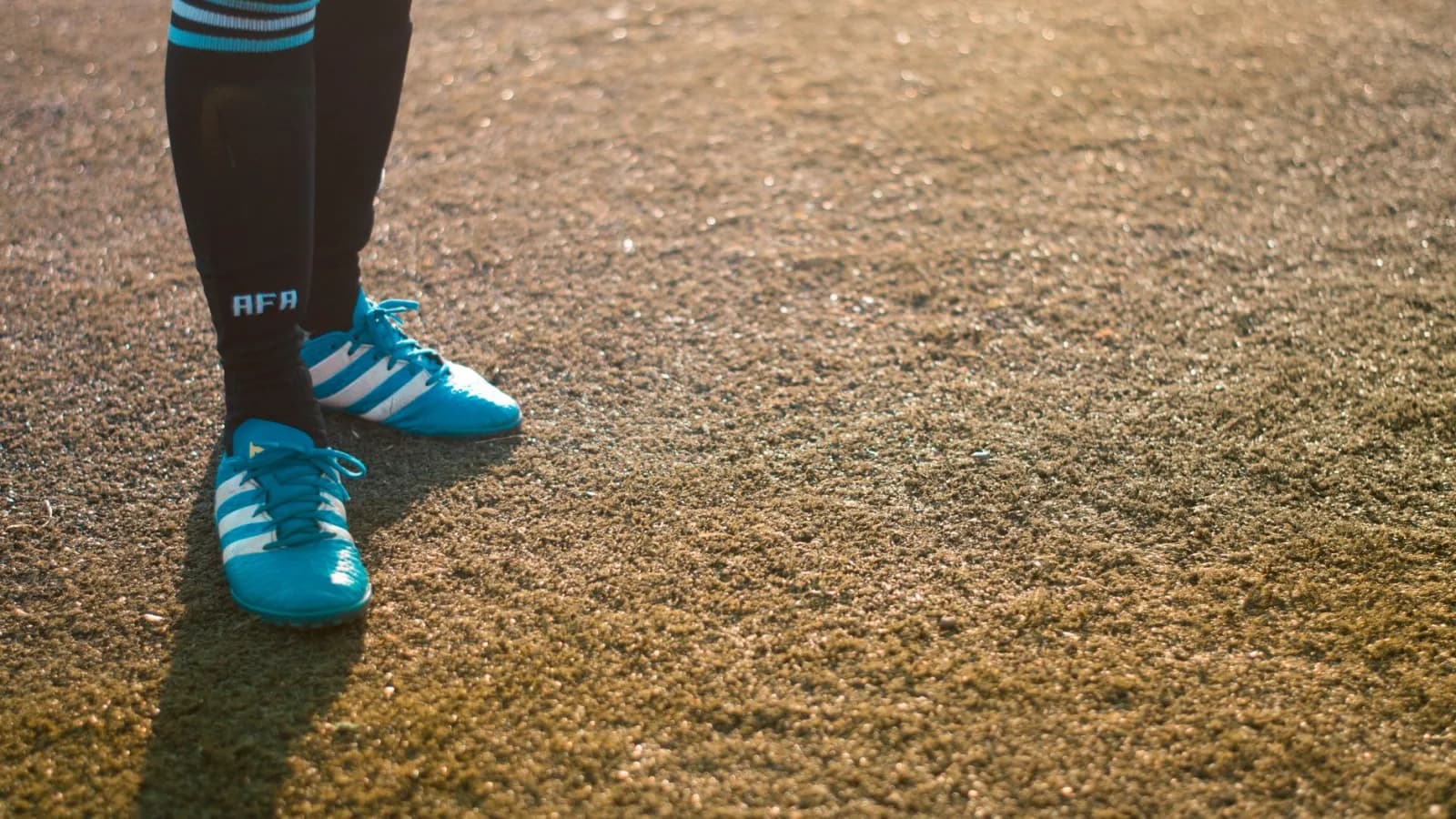 Këmbët e një futbollisti me atlete blu dhe çorape të zeza në një fushë bari.