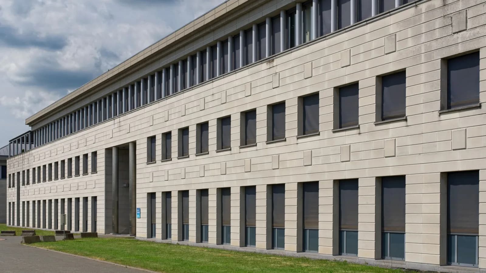 Fasada e një ndërtese moderne administrative me dritare të shumta dhe hapësirë të gjelbër përpara.
